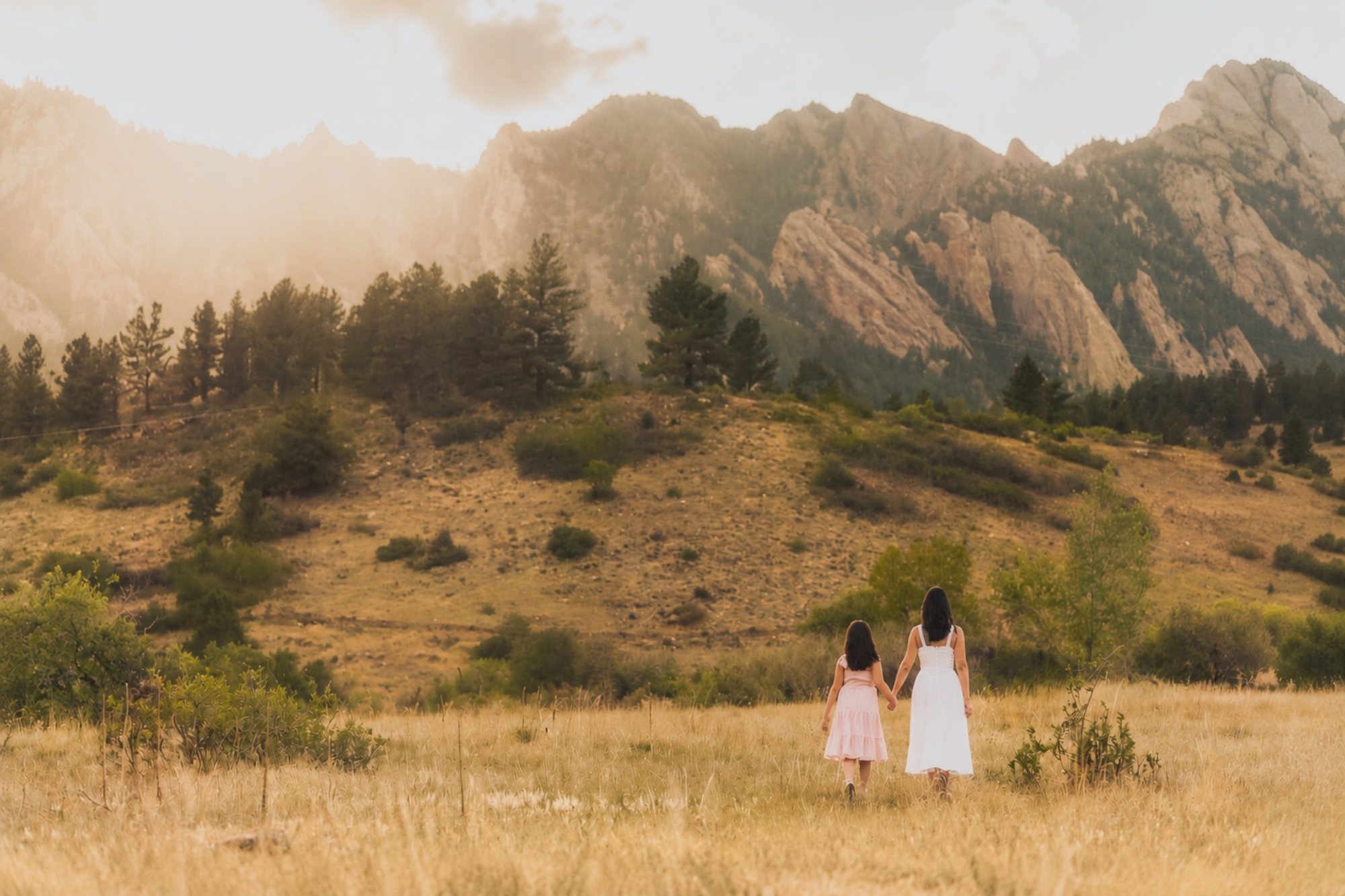 A mother and daughter walking hand-in-hand through a golden grass field, the Flatirons rising behind them at golden hour.